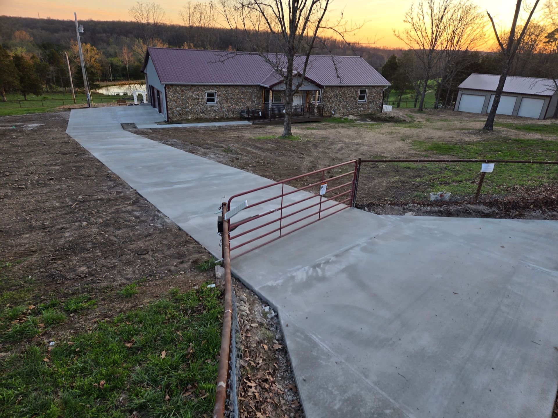 Concrete driveway leading to a stone house at sunset, with a rusty gate in the foreground.