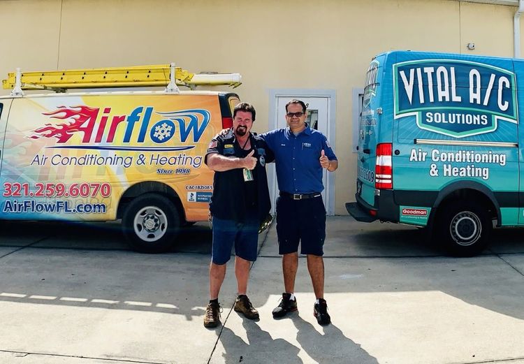 Two men standing between Airflow and Vital A/C service vans in a sunny parking lot.