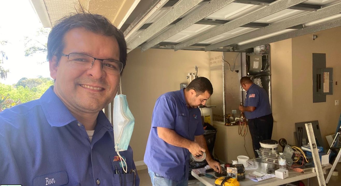 Three men in blue shirts working in a garage, with tools and parts on a table.