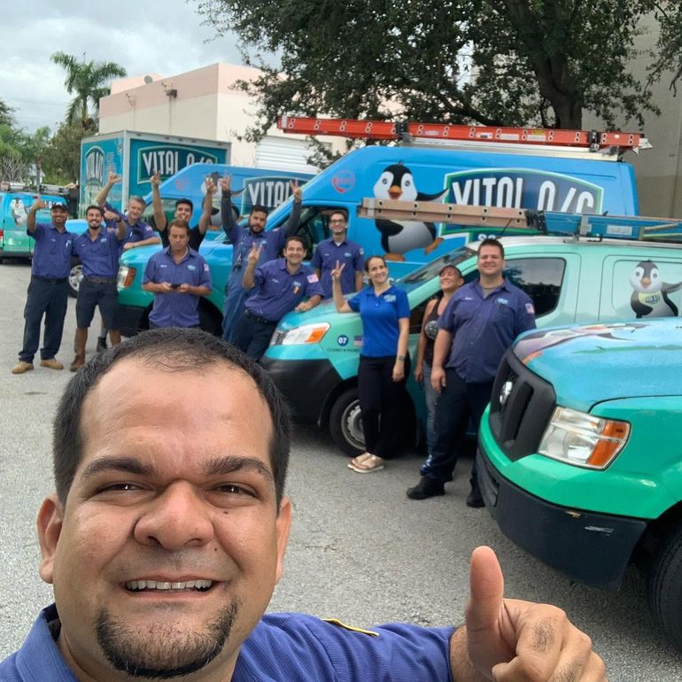 Group of utility workers posing with blue service trucks, one man smiling in the foreground with a thumbs-up.