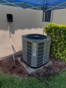 Outdoor air conditioner unit beside a beige house wall, under a blue canopy, near a green shrub.
