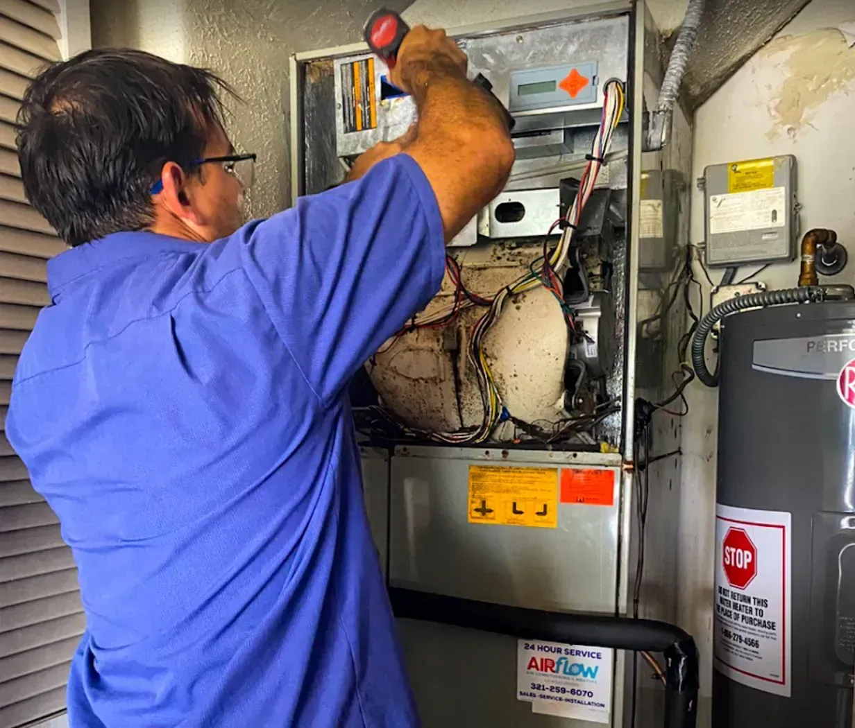 Technician in blue shirt inspecting HVAC unit in a utility room with exposed wiring and pipes