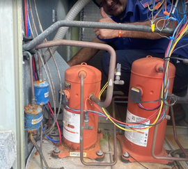Two orange industrial compressors with colored wires and pipes in a maintenance area, worker partially visible above