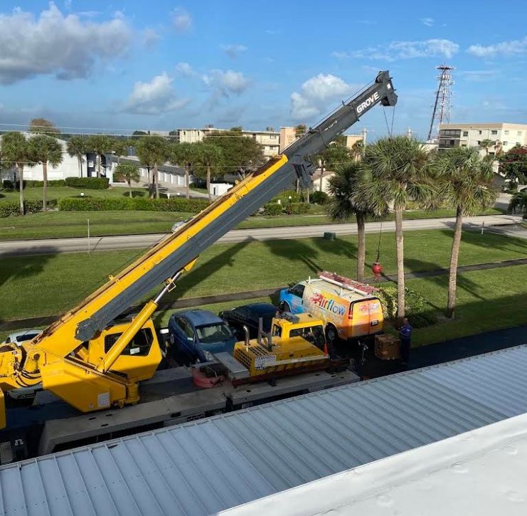 Yellow crane lifting equipment beside parked cars in a sunny roadside area with palm trees