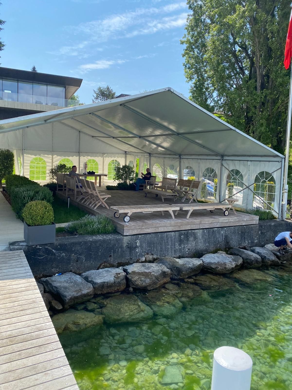 Tente blanche avec des tables installées sur une terrasse en bois au bord d'un lac, avec des arbres et un bâtiment en arrière-plan.