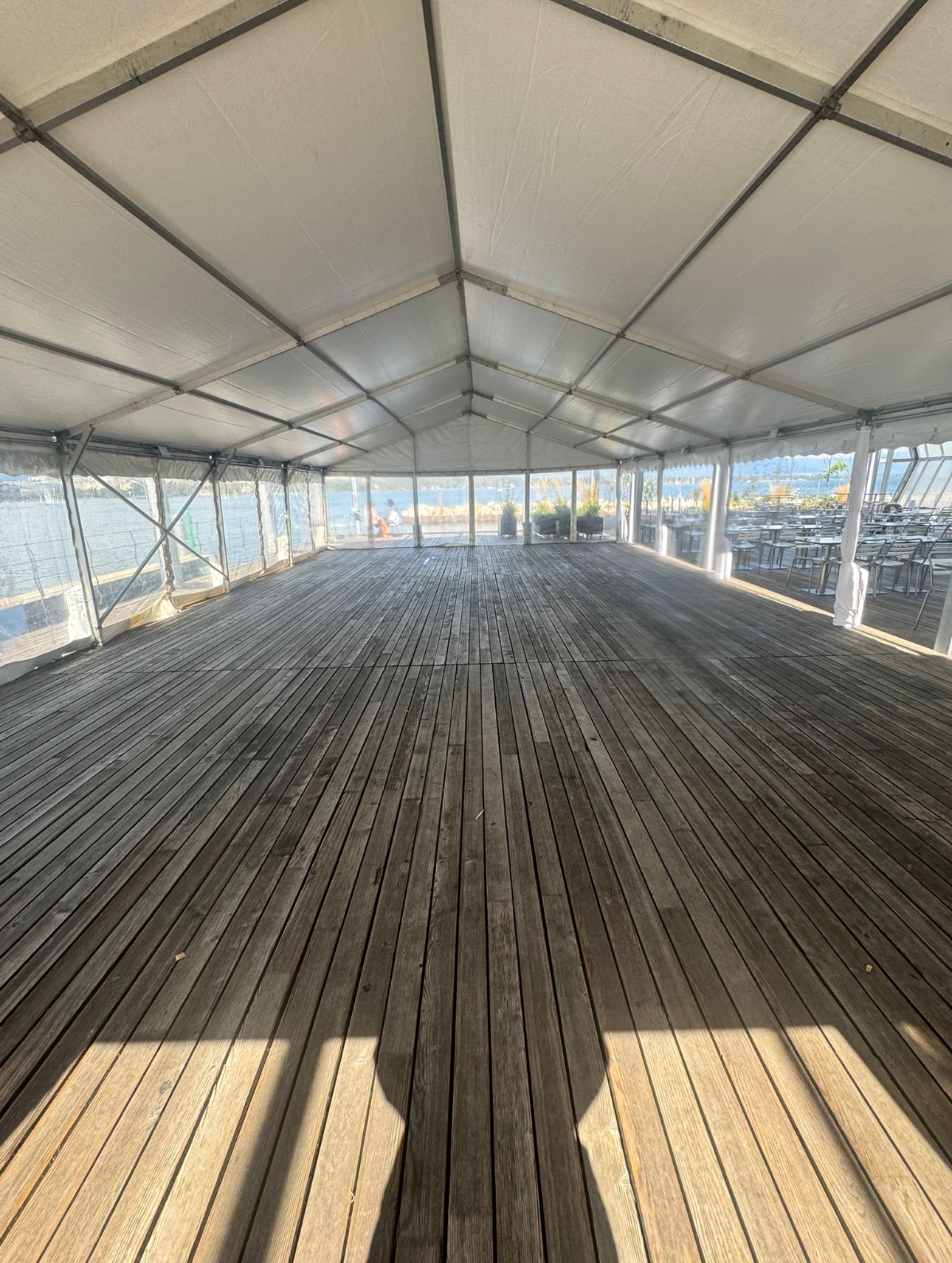 Intérieur d'une grande tente avec plancher en bois, panneaux latéraux transparents et toit blanc ; vue vers l'extérieur.
