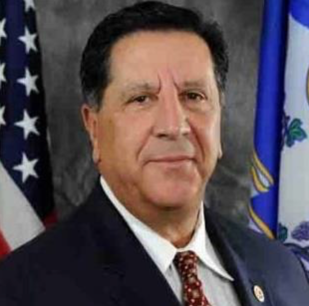 Man in suit, smiling, with US and state flags in background.