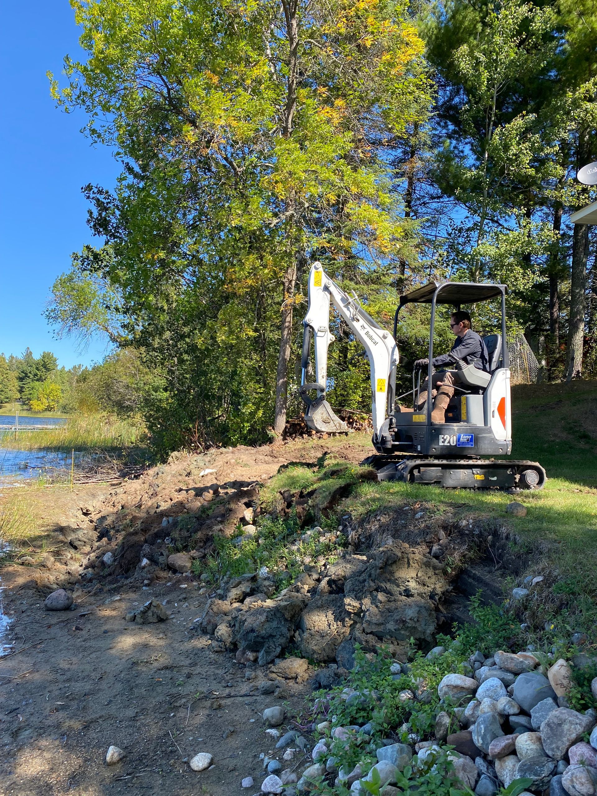 A man is driving a small excavator next to a body of water.