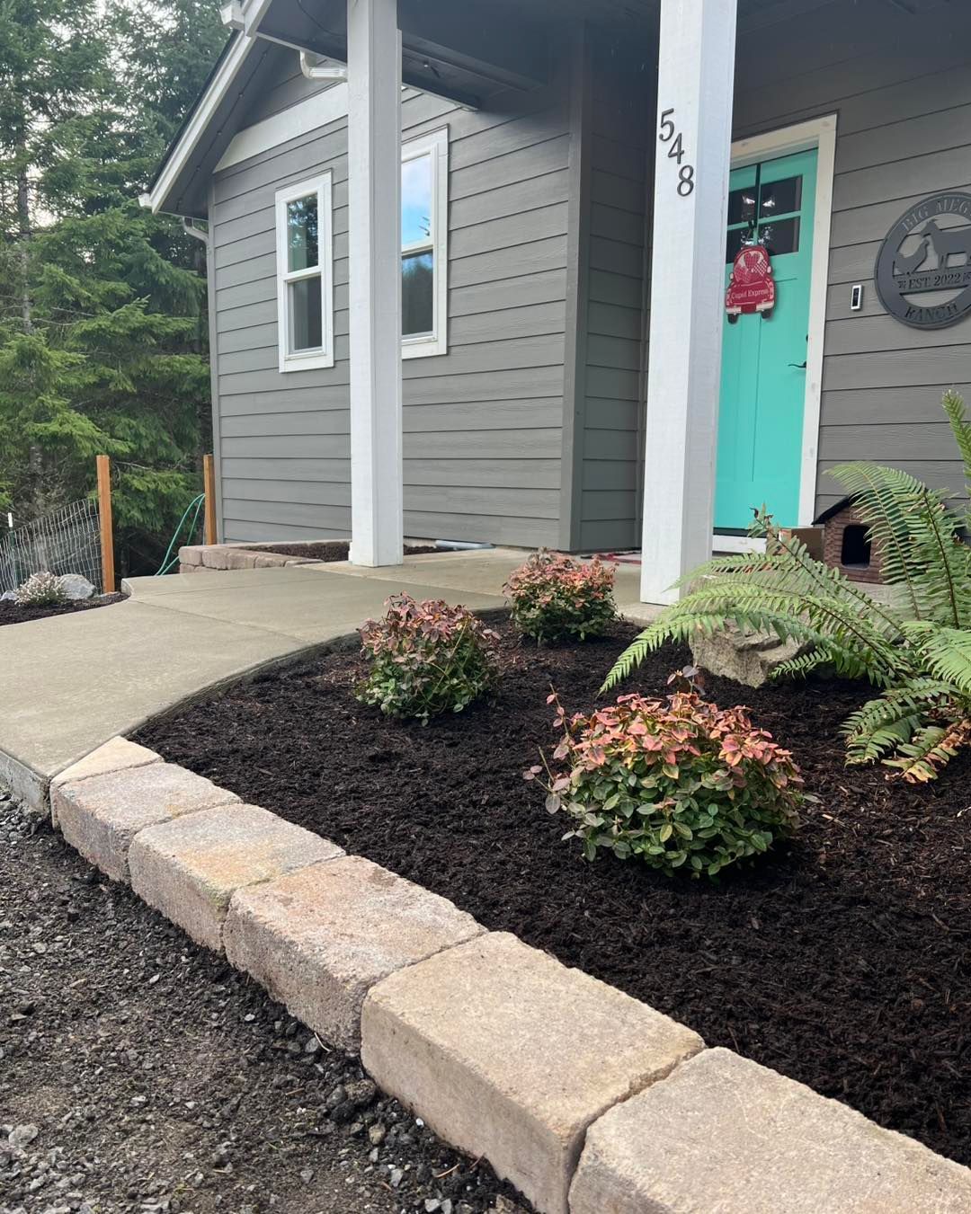 A house entrance featuring a teal door, gray siding, and a landscaped garden bed with mulch and shrubs behind stone pavers.