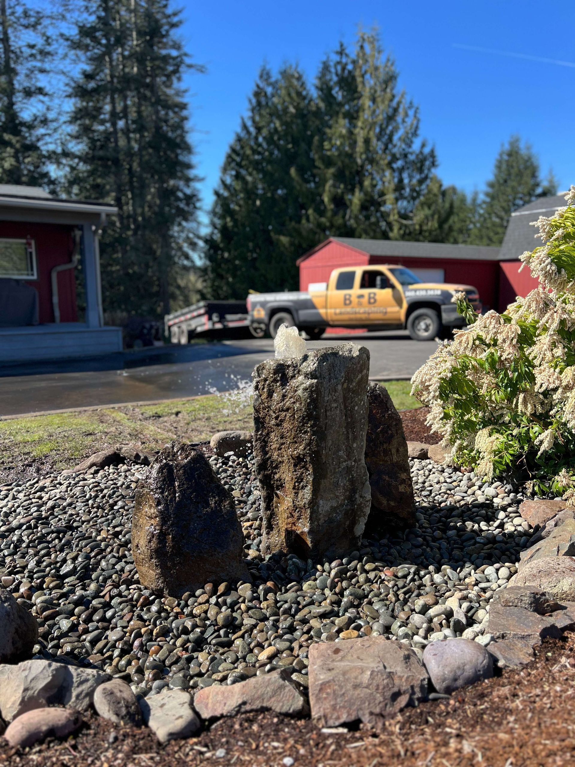 Garden mulch mound beside a driveway, with red buildings and a yellow truck in the background.