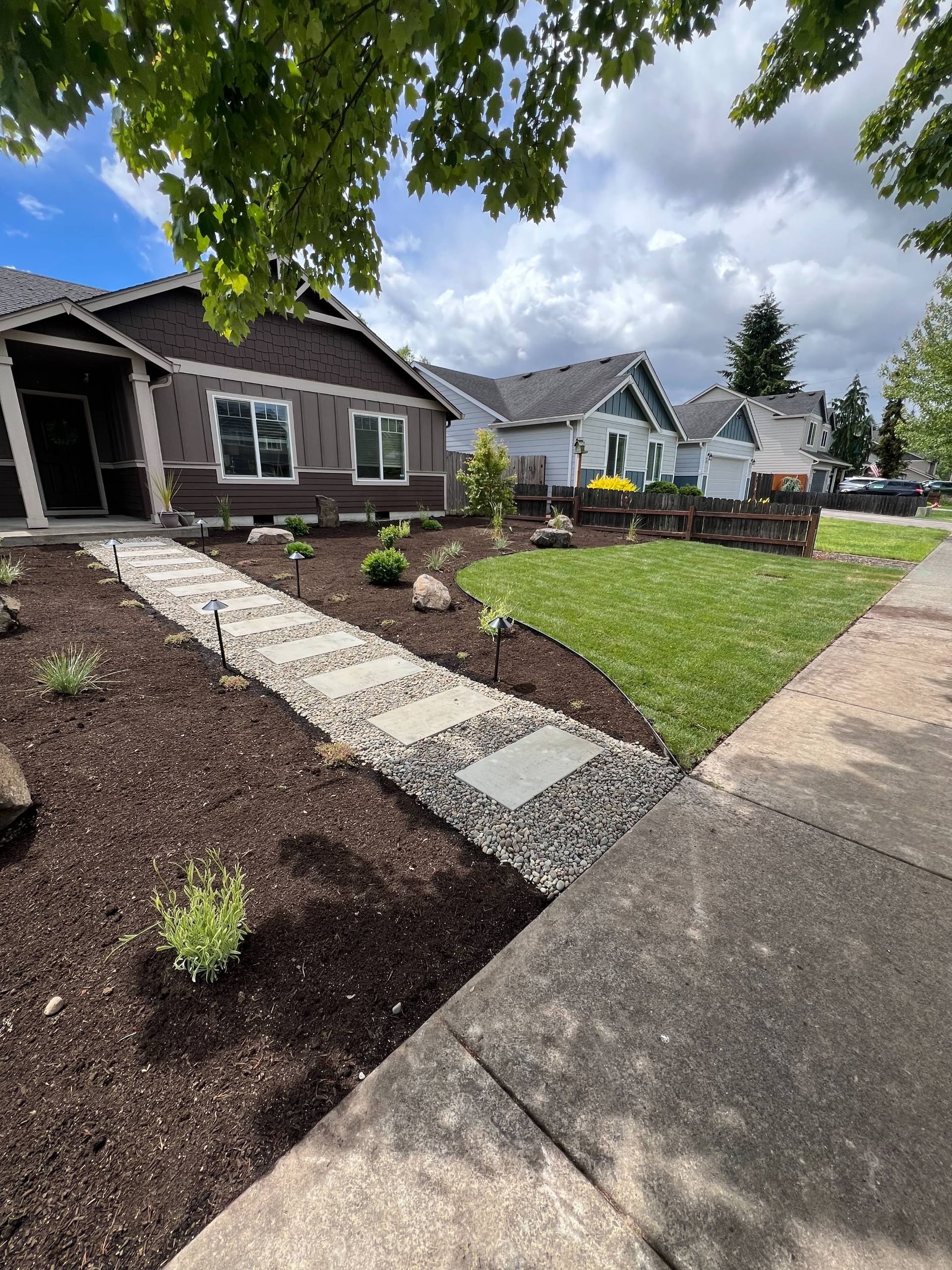 Landscaped suburban homes with stone walkway, mulch beds, and green lawn under cloudy sky