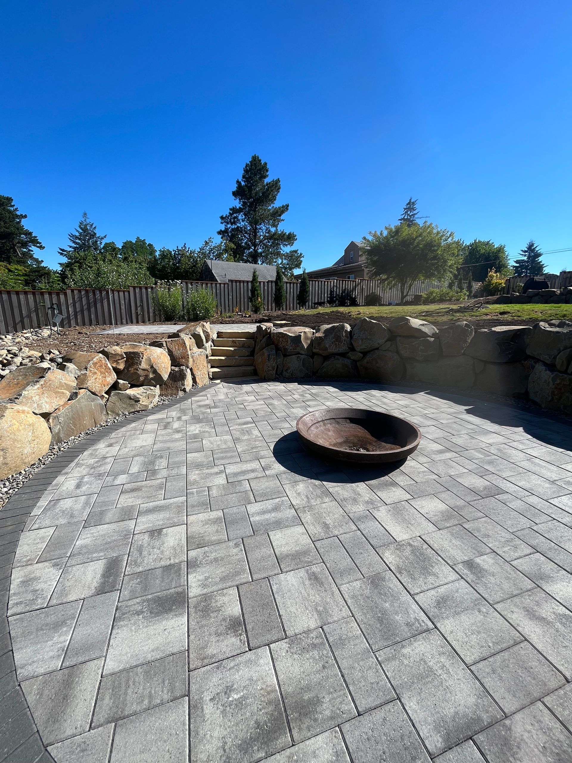 Circular stone patio with a fire pit, surrounded by rocks and greenery, under a blue sky.