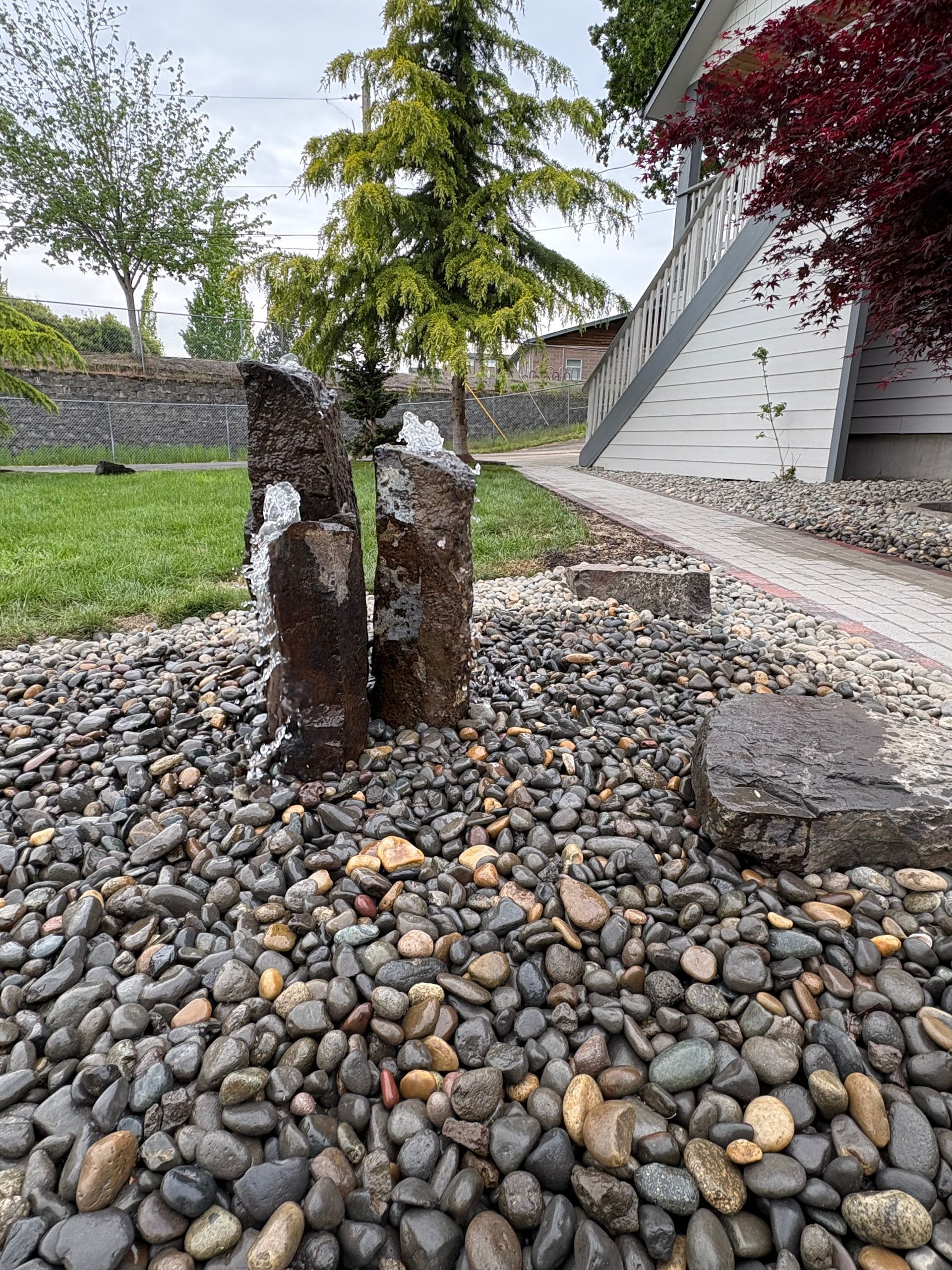 A stone fountain with water cascading over dark rocks surrounded by pebbles, in a garden.