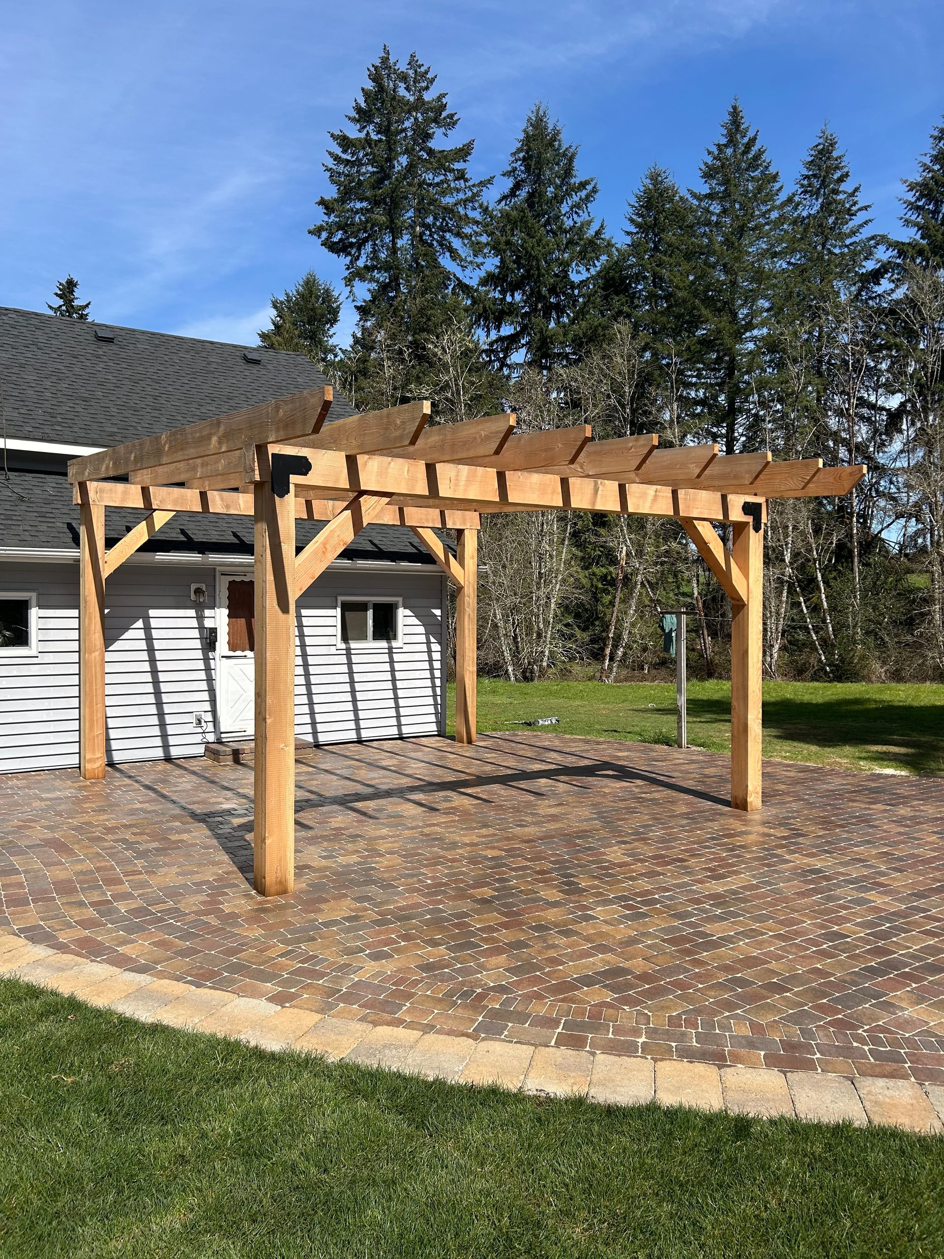 Wooden pergola on a brick patio, in front of a house with trees in the background on a sunny day.