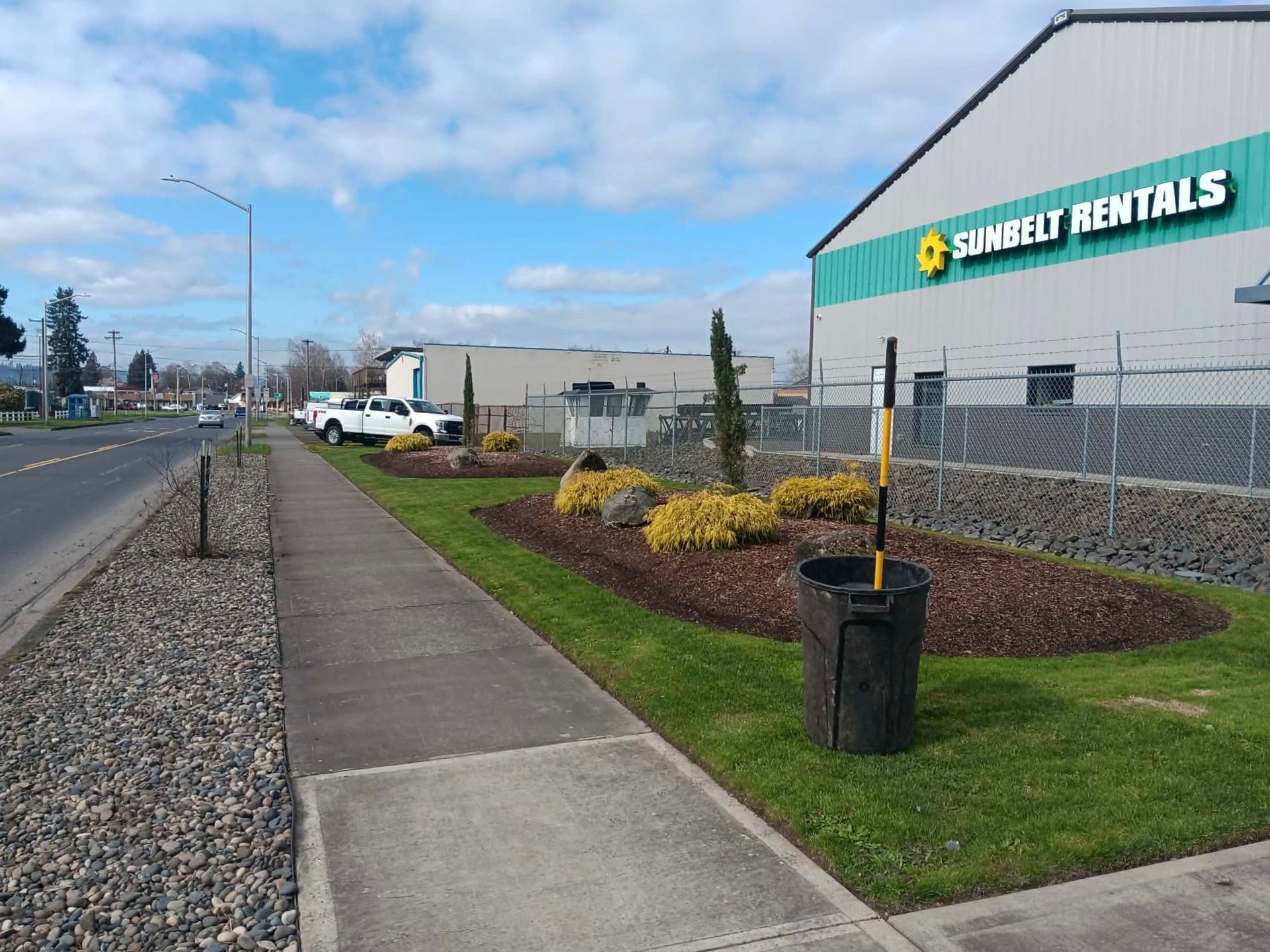 A sidewalk next to a landscaped grass strip, a gravel shoulder, and a Sunbelt Rentals building under a cloudy blue sky.