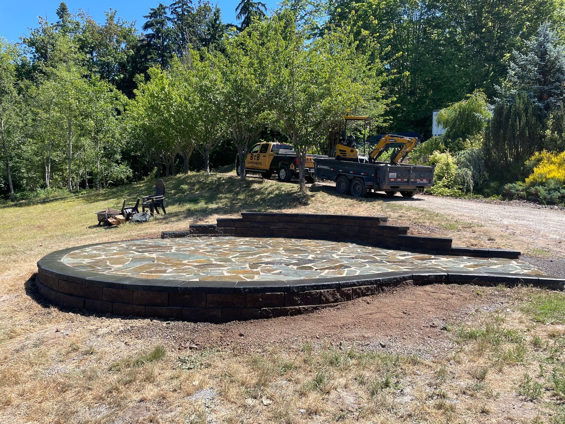 Stone patio with tiered walls under construction; machinery and trees in background.