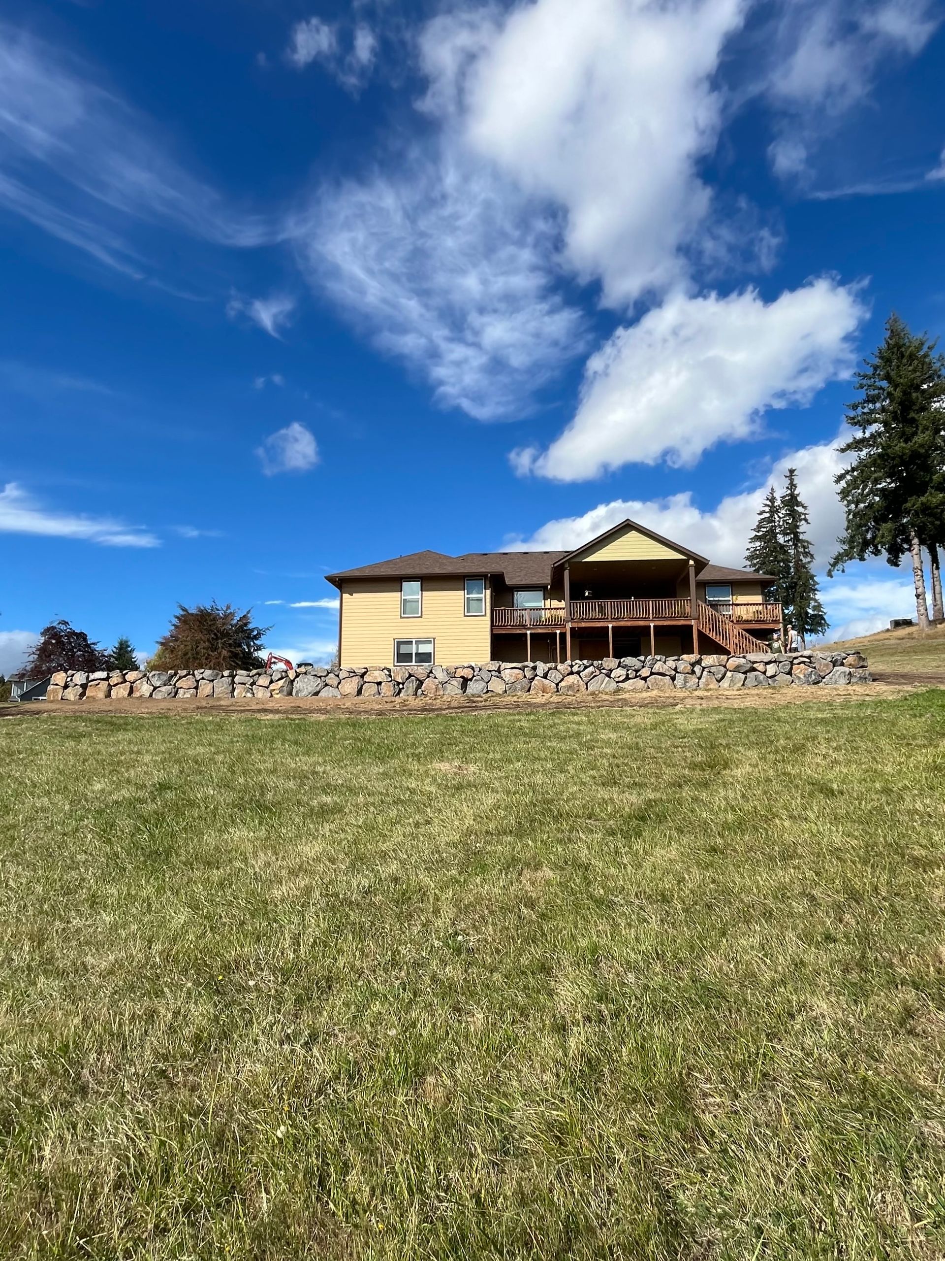 A yellow house with a porch sits on a grassy hill under a blue sky with clouds.
