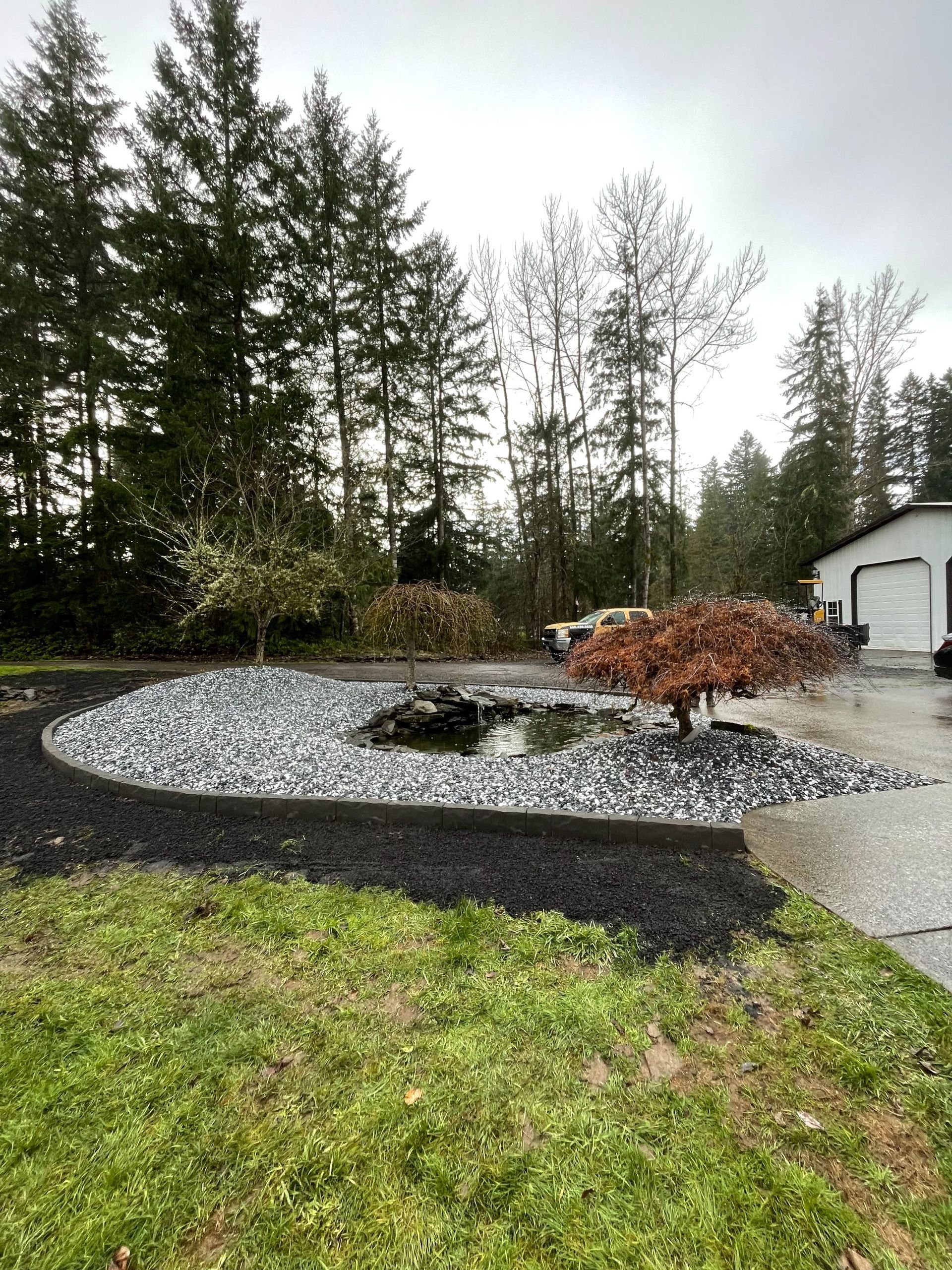 A landscaped garden bed with a small pond, surrounded by white pebbles and black mulch, next to a house and pine trees.