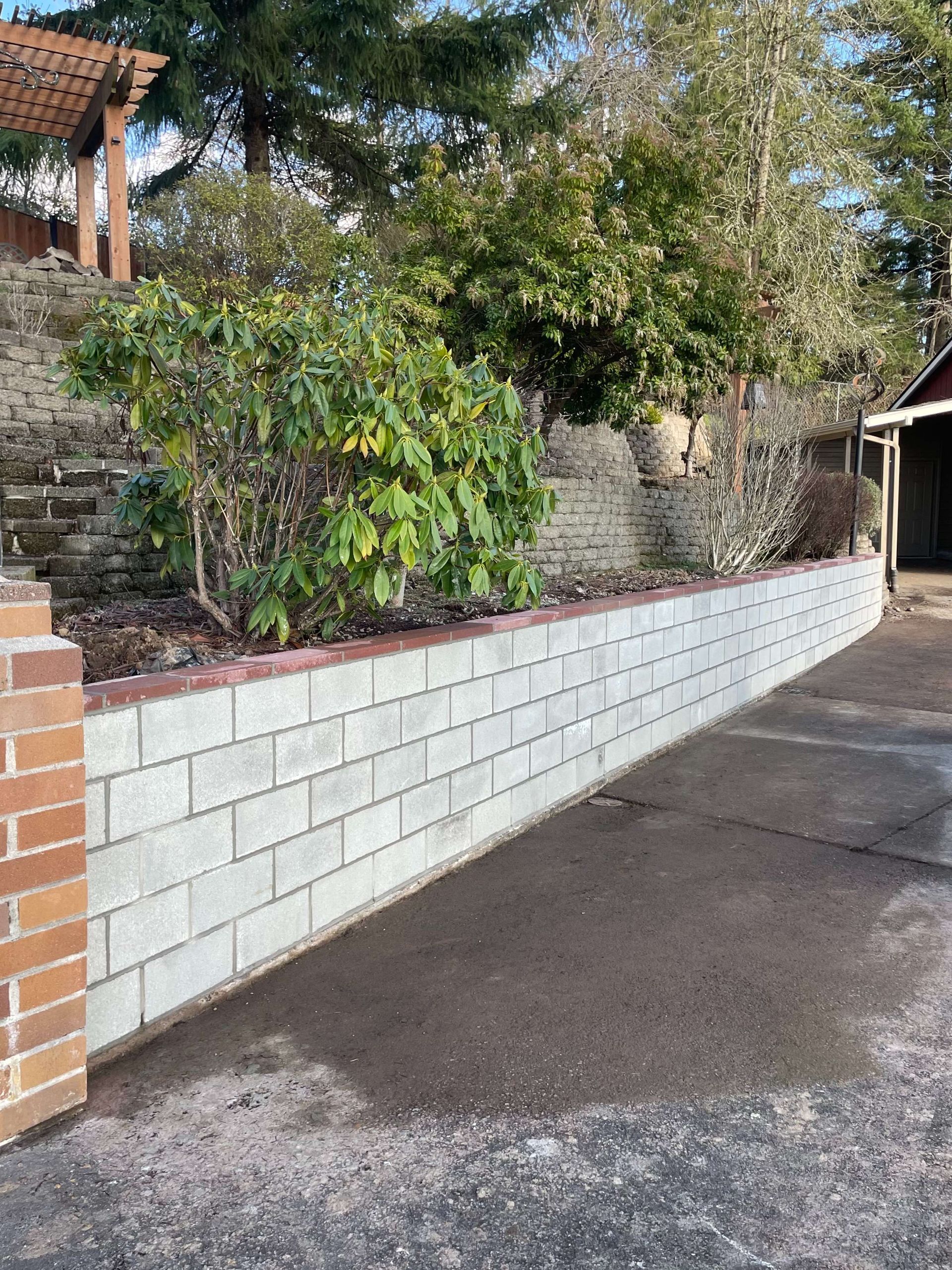 A light-colored concrete block retaining wall lines a driveway next to a garden with shrubs and trees.