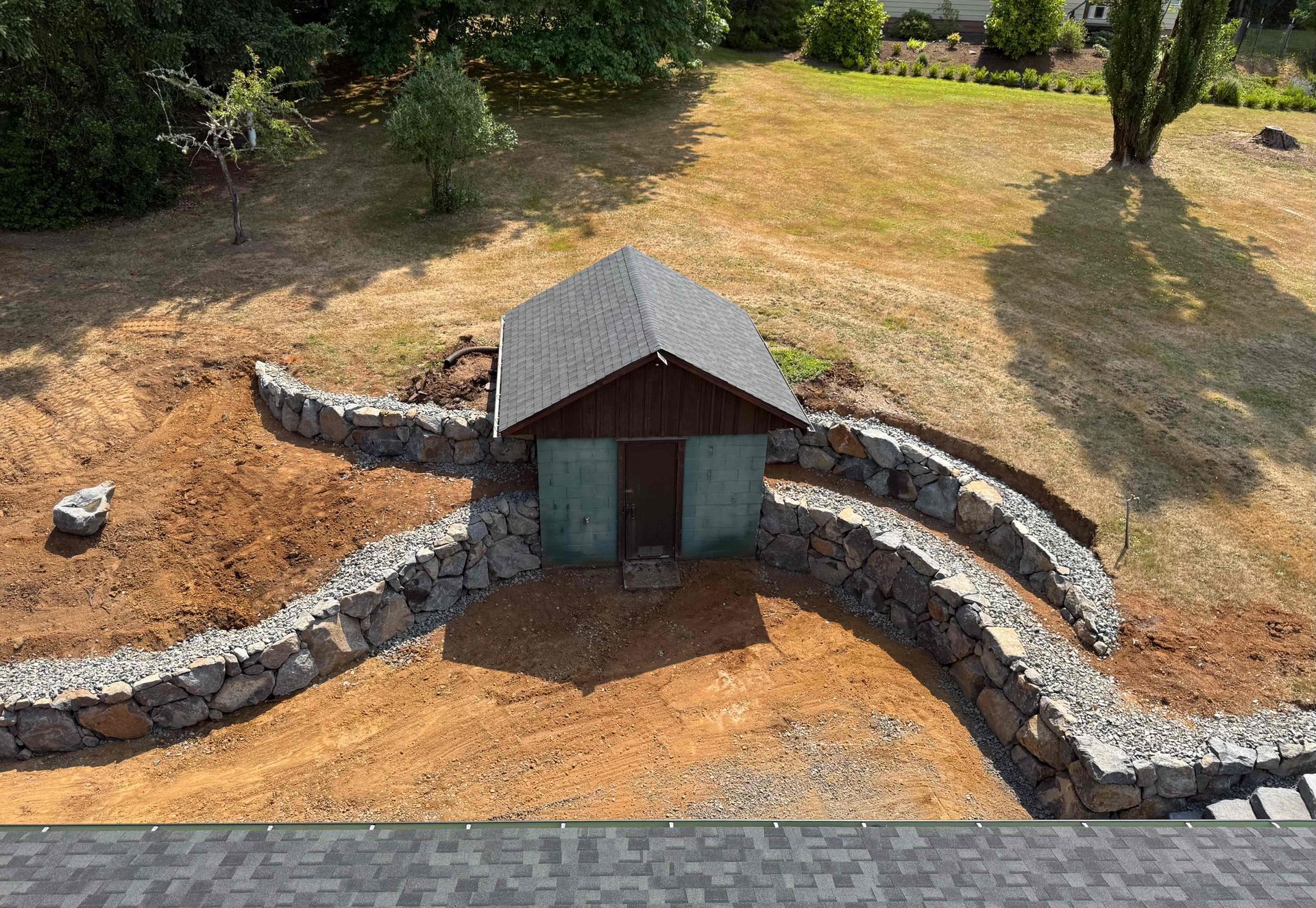 Small stone-walled garden structure with a dark roof, set in a dry grassy yard.