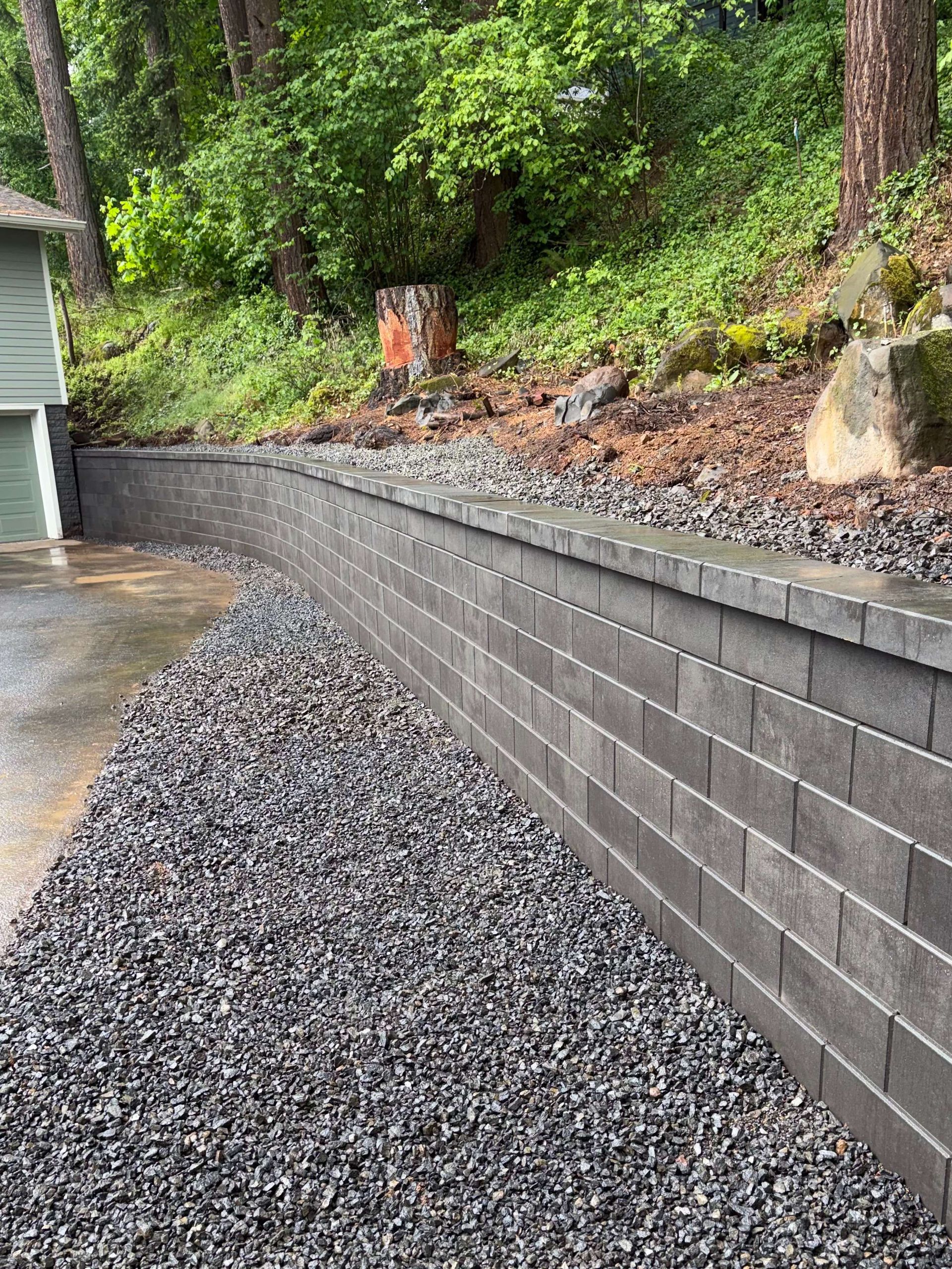 Gravel-filled retaining wall beside a driveway and wooded slope near a house