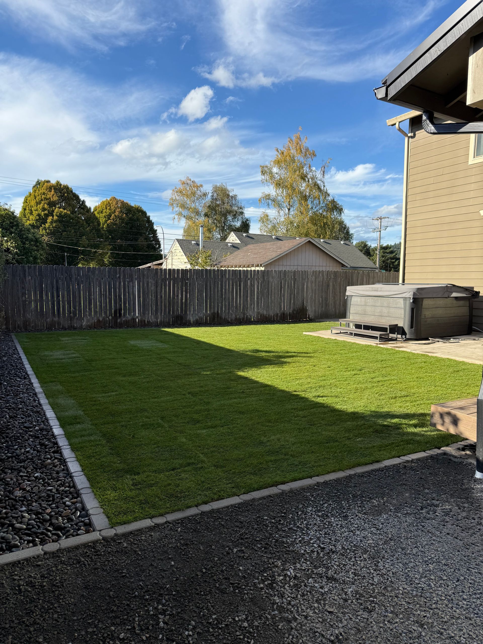 Backyard with green lawn, wooden fence, blue sky, and patio area beside a house
