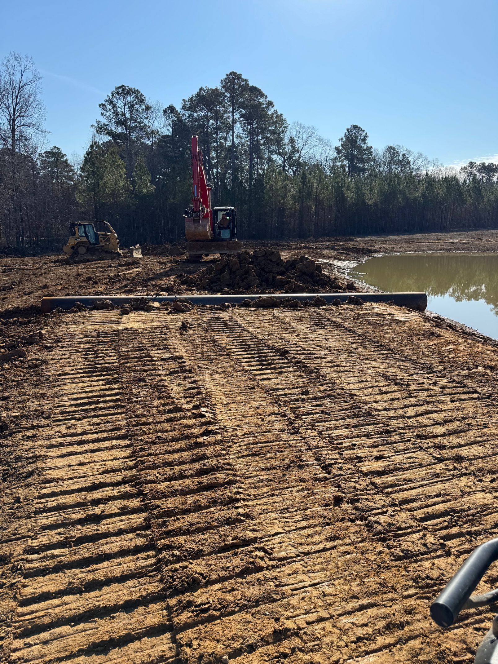 Construction site with a red excavator on a dirt embankment beside muddy water and trees under a clear sky