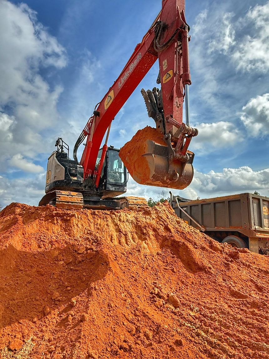 A red excavator on a large mound of orange dirt scoops a bucket full of soil near a dump truck under a blue, cloudy sky.