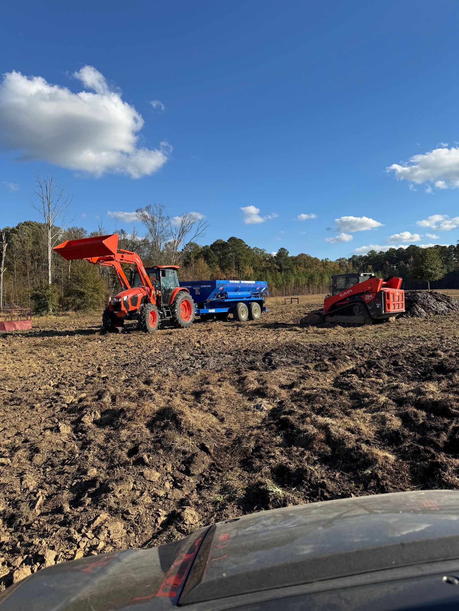 Orange tractor with blue trailer and red machine in a muddy field under a blue sky