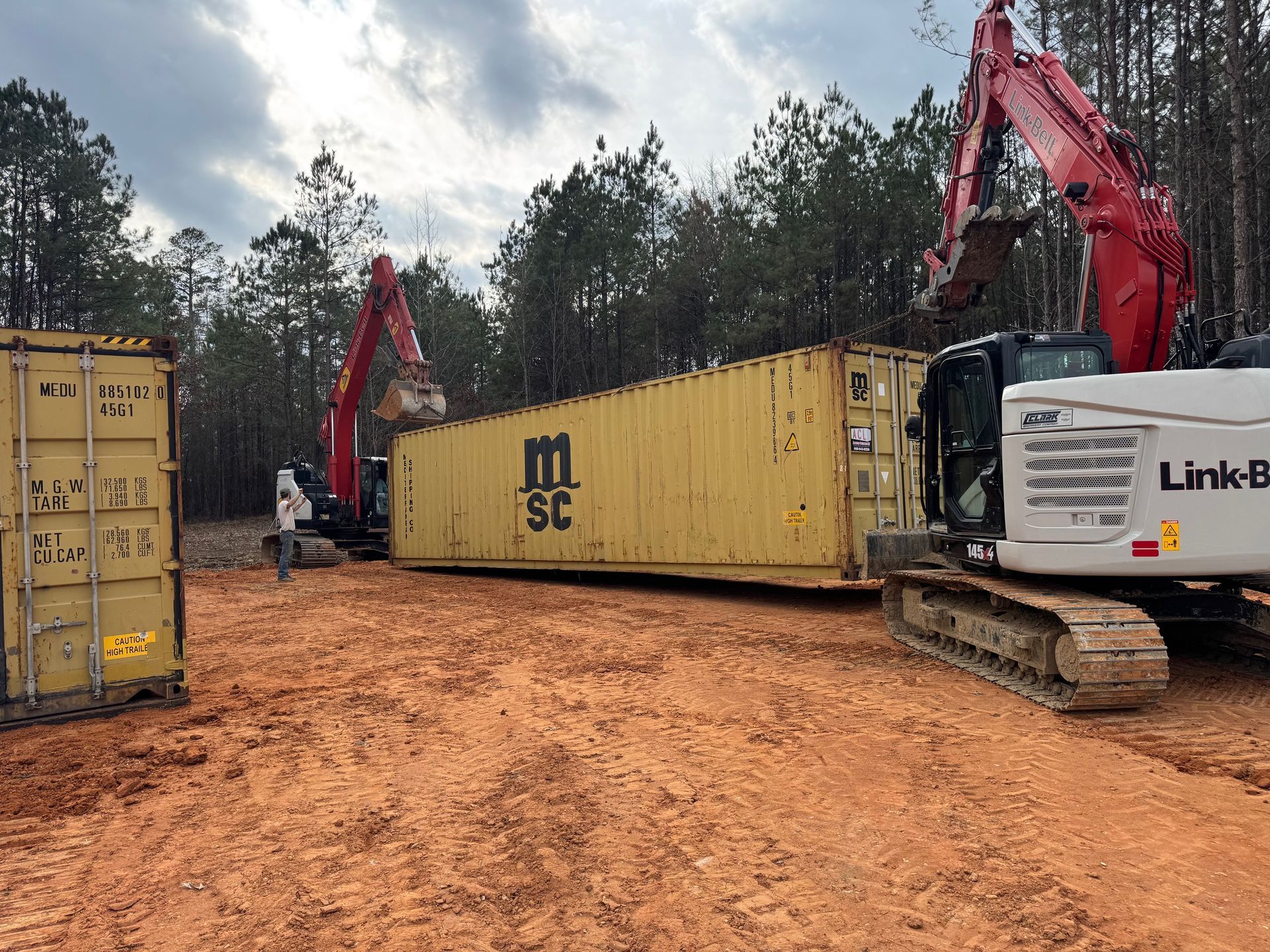 Two excavators loading yellow shipping containers on a muddy construction site in a wooded area