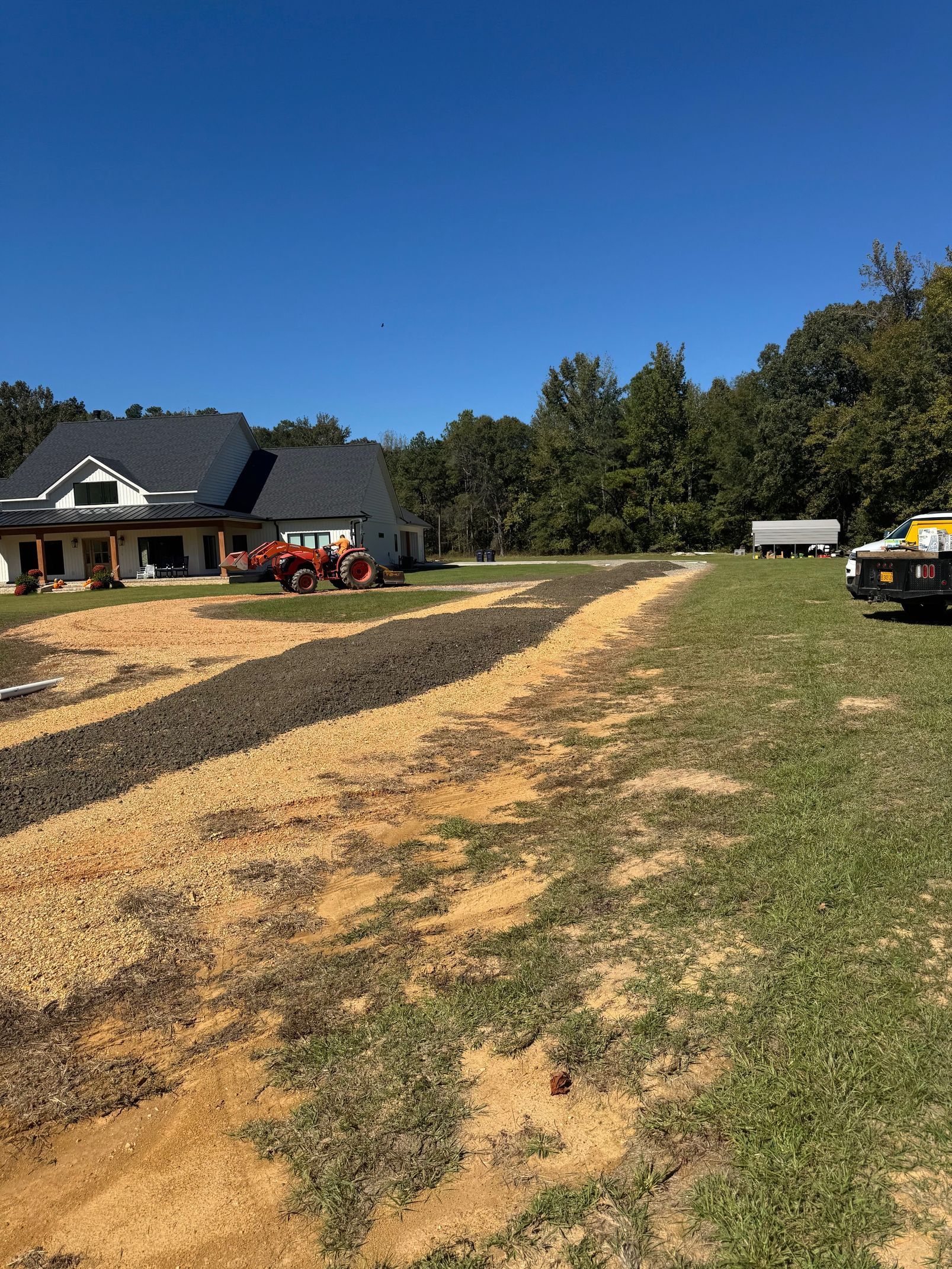 Rural dirt driveway beside a house, with parked vehicles and trees under a clear blue sky.