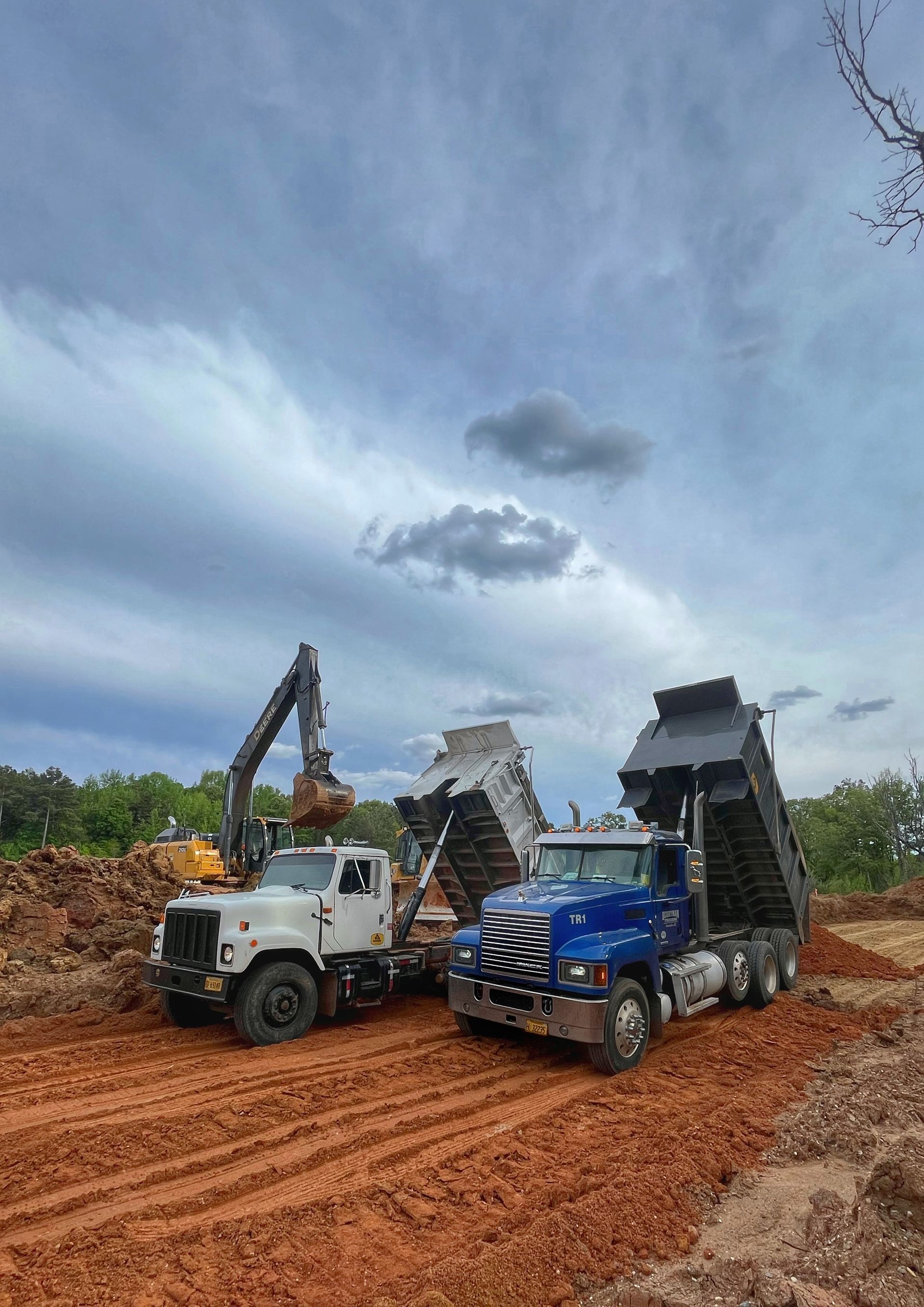 Two dump trucks on a muddy dirt road under a cloudy sky, unloading debris at a worksite