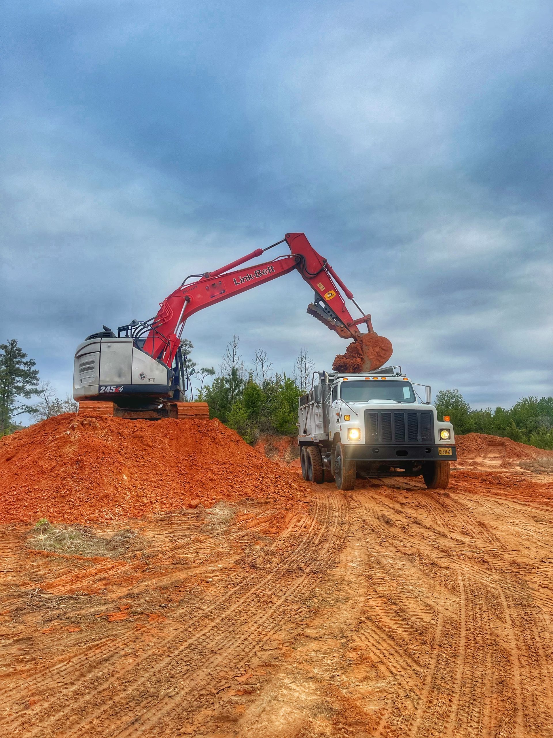 Excavator loading orange dirt into a dump truck at a construction site under a cloudy sky