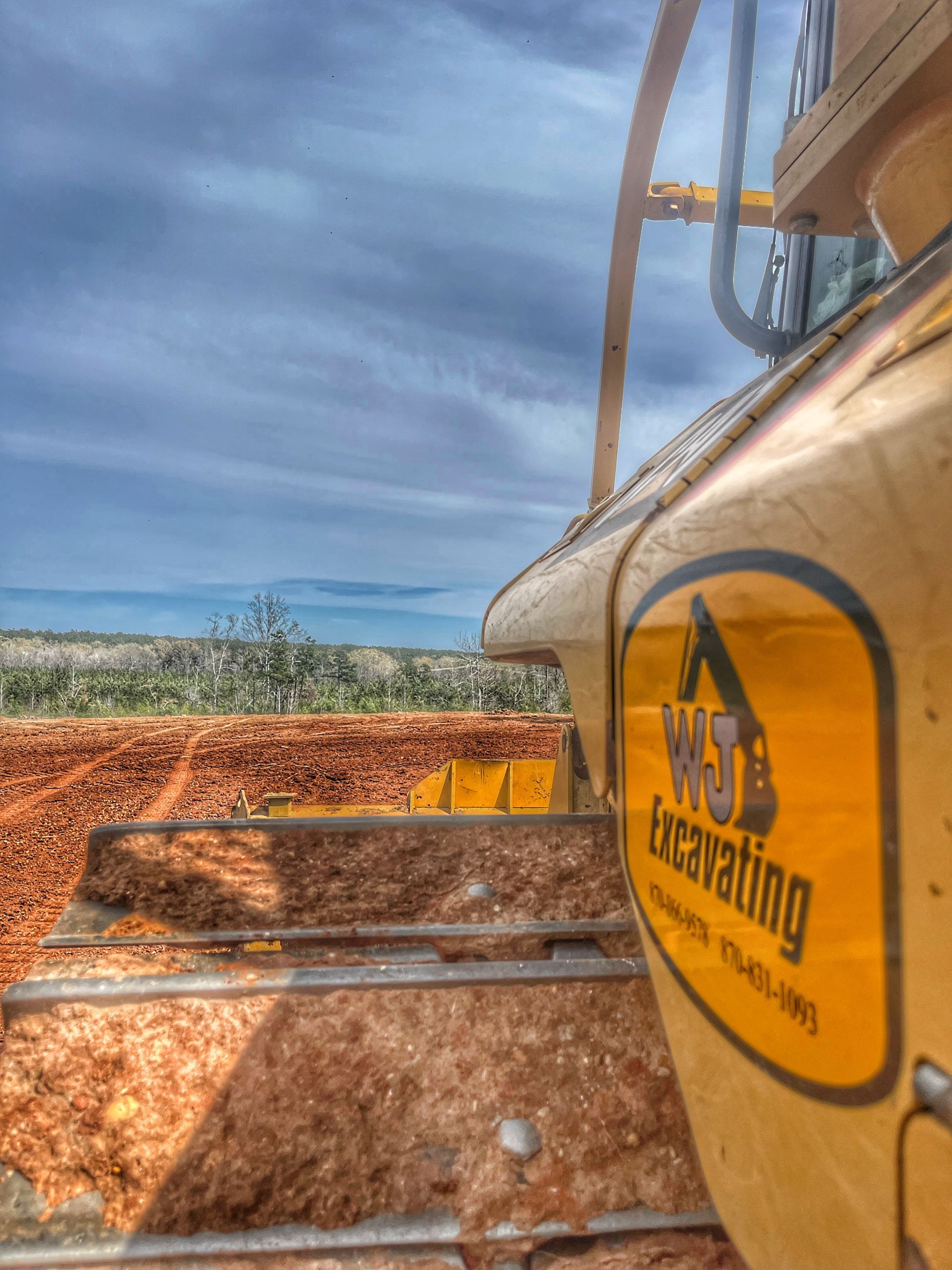 Yellow excavator arm and cab beside a red dirt work site under a cloudy sky.