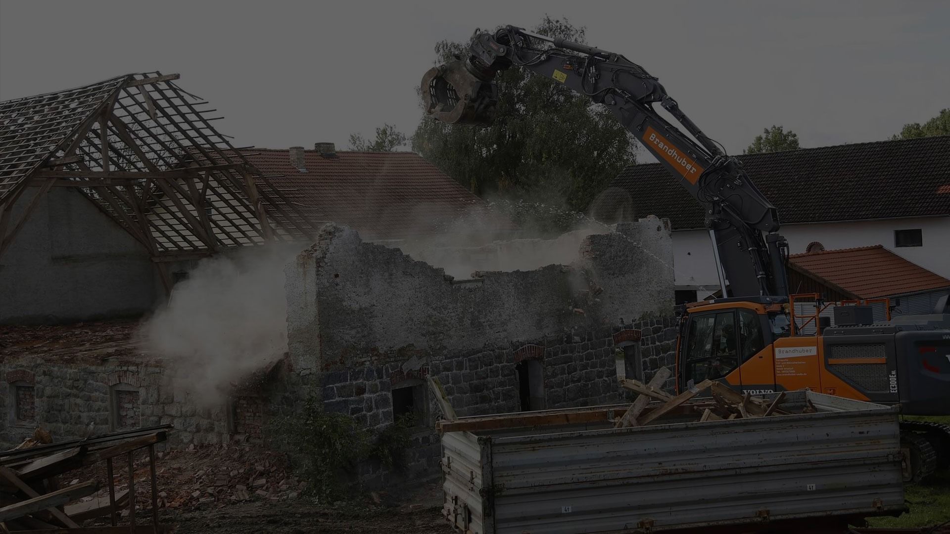 An excavator demolishes a stone wall, creating a cloud of dust near older buildings with exposed roof rafters.