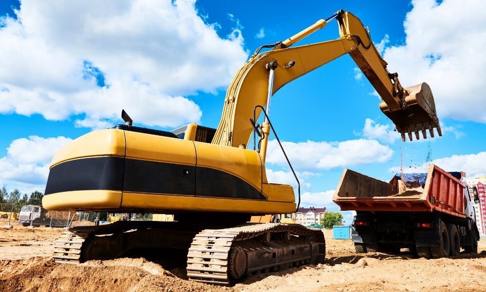 A yellow excavator loading soil into a dump truck at a construction site against a bright blue, cloudy sky.