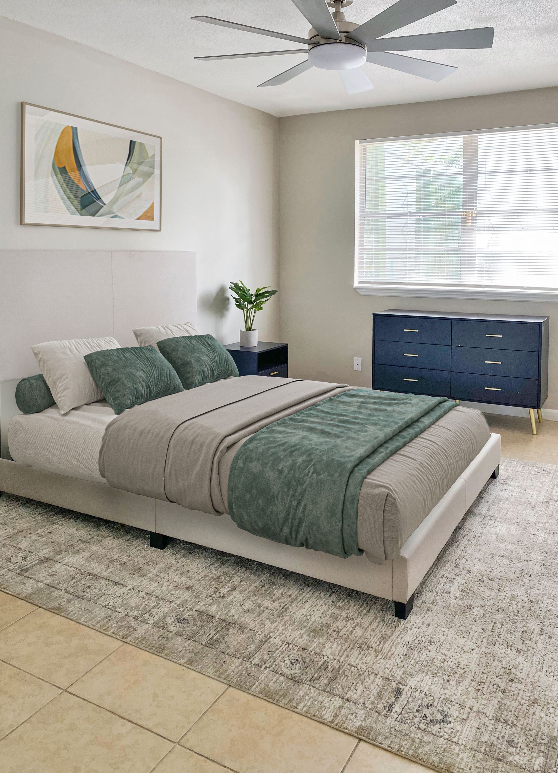 Bedroom with a bed, blue dresser, artwork, rug, and window with blinds at Caroline Village in Jacksonville, FL.