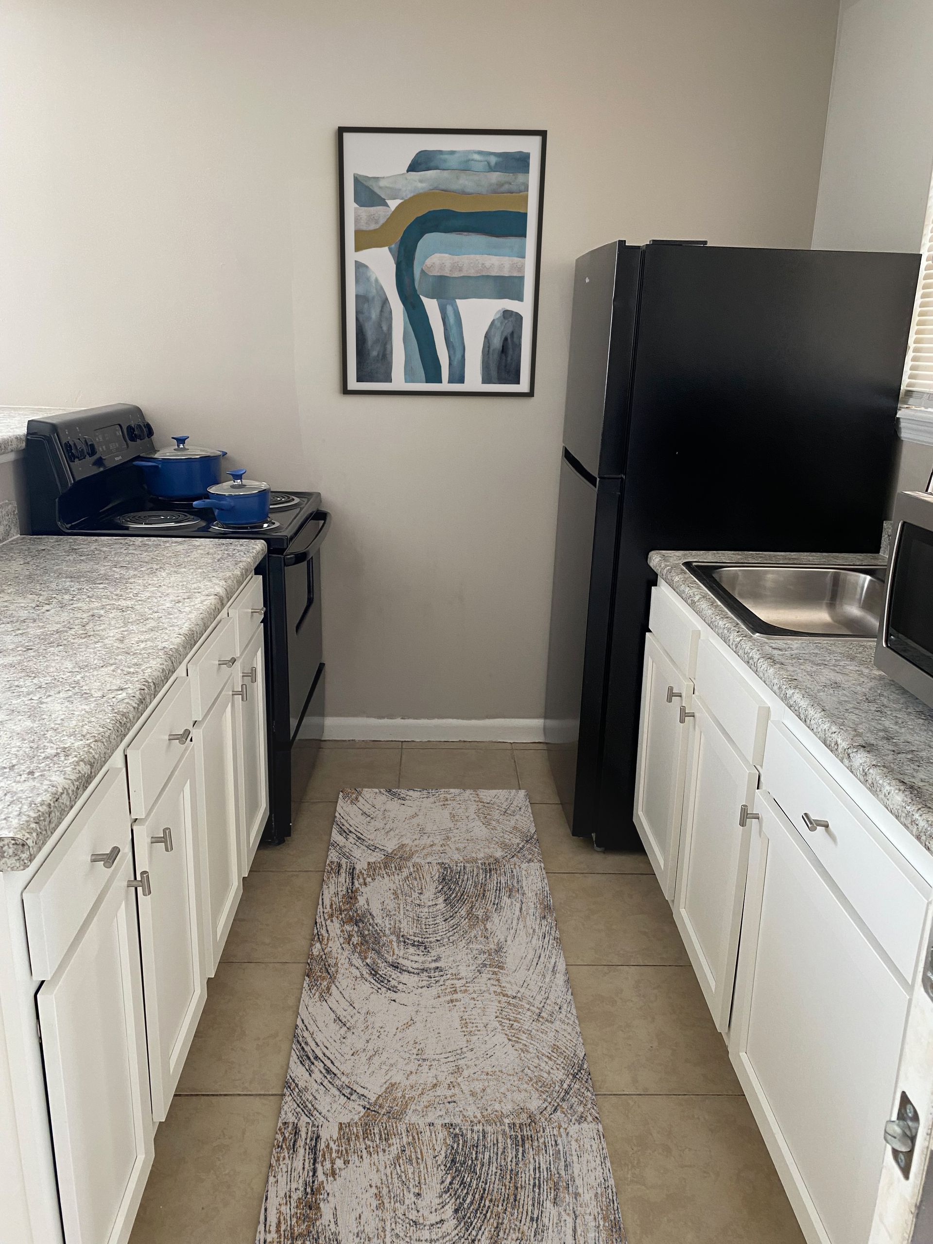 Narrow kitchen with white cabinets, black appliances, and a rug. Artwork hangs between the refrigerator and stove at Caroline Village in Jacksonville, FL.