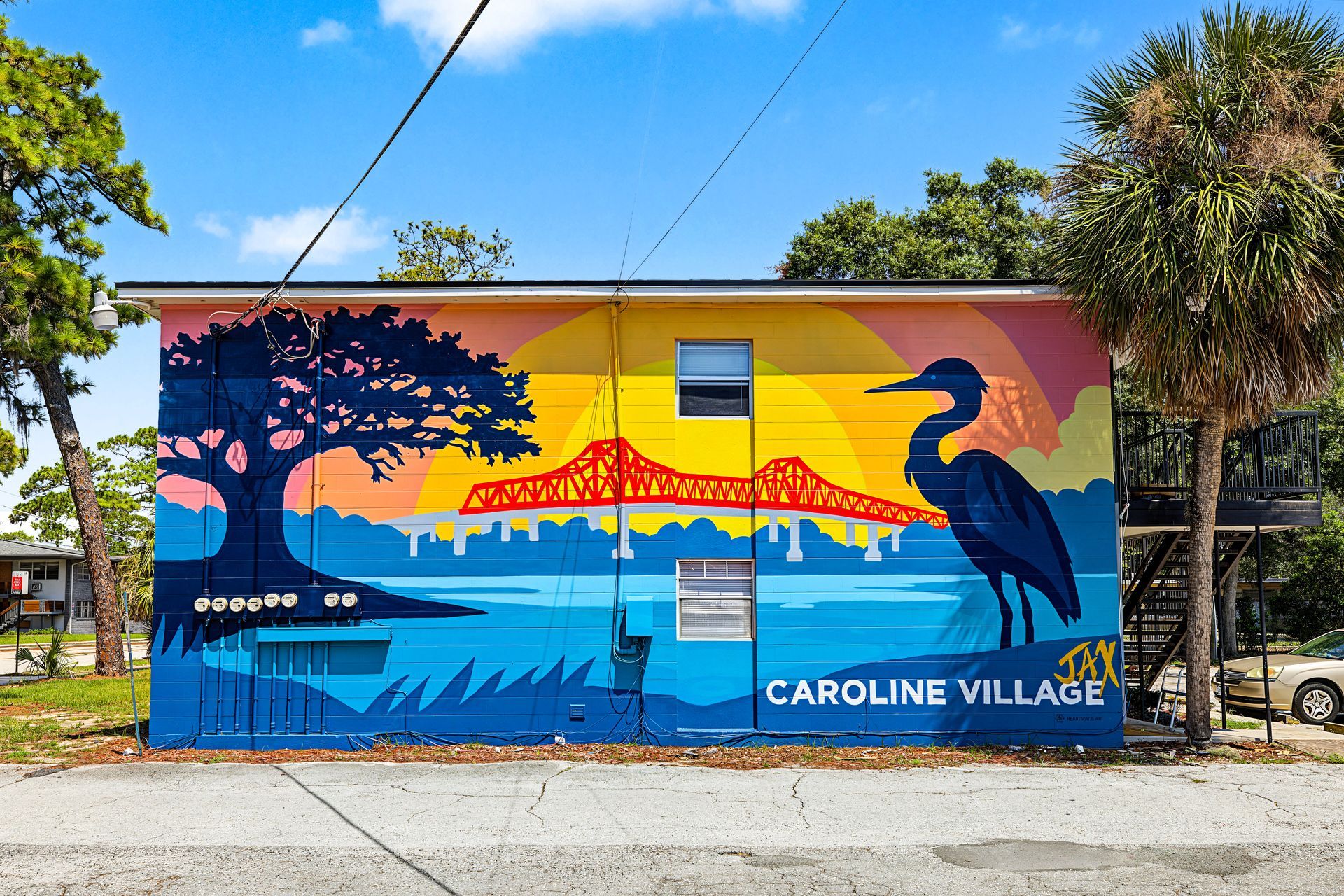 Colorful mural of a tree, sunset, water, and a bird on a building labeled “Caroline Village” at Caroline Village in Jacksonville, FL.