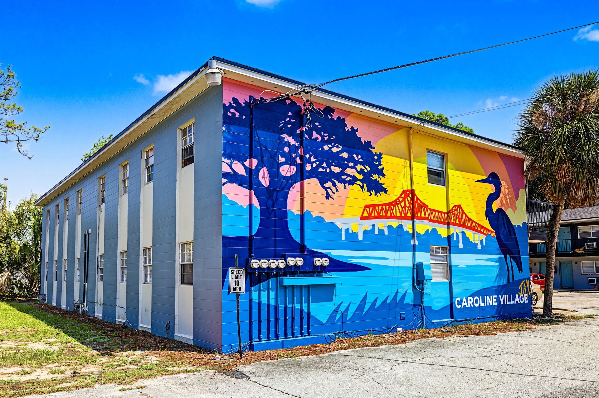 Colorful mural on a building wall with a sunset, tree silhouette, and blue ocean scene under a clear sky at Caroline Village in Jacksonville, FL.