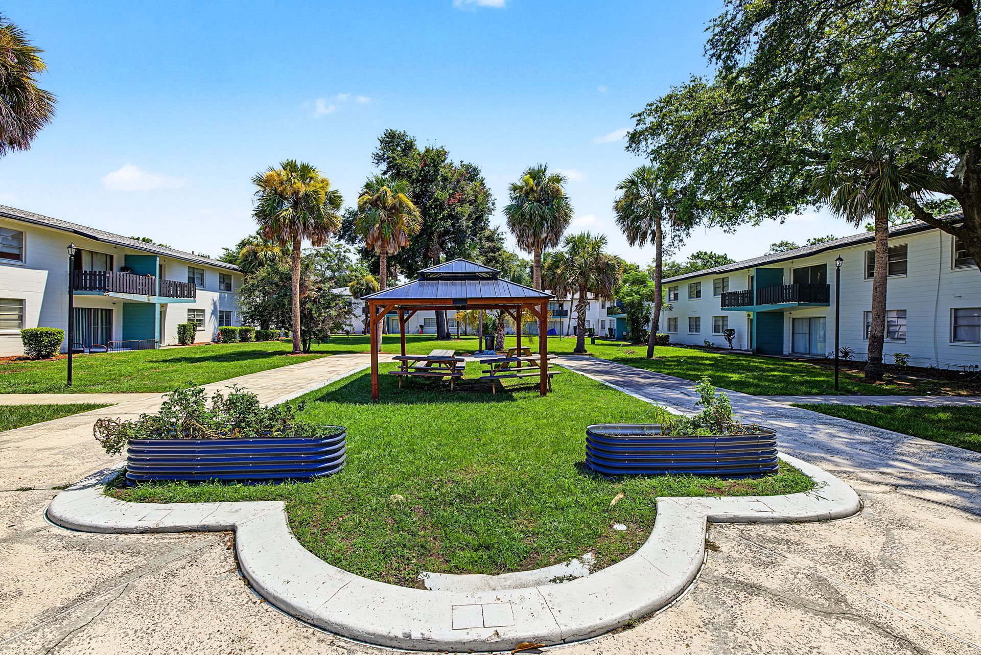 Courtyard with central gazebo, palm trees, green lawn, and white apartment buildings under a blue sky at Caroline Village in Jacksonville, FL.