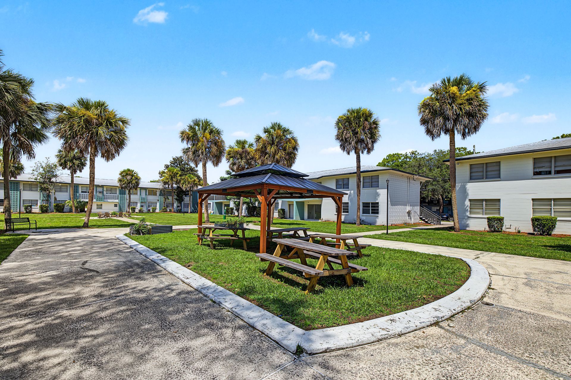Courtyard with picnic tables, palm trees, and apartment buildings under a blue sky at Caroline Village in Jacksonville, FL.
