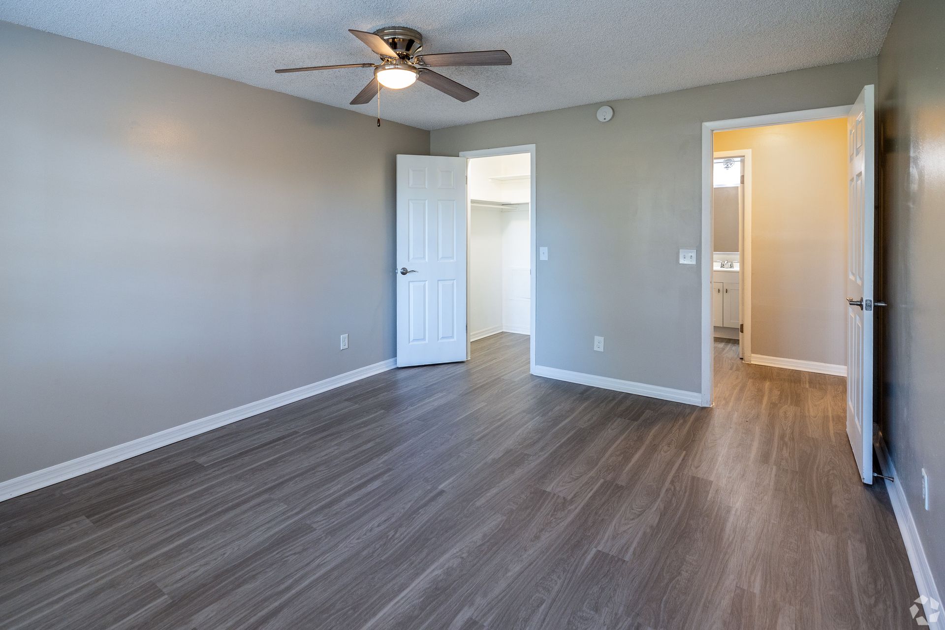 Empty bedroom with gray walls, wood-look flooring, and three doorways at Caroline Village in Jacksonville, FL.