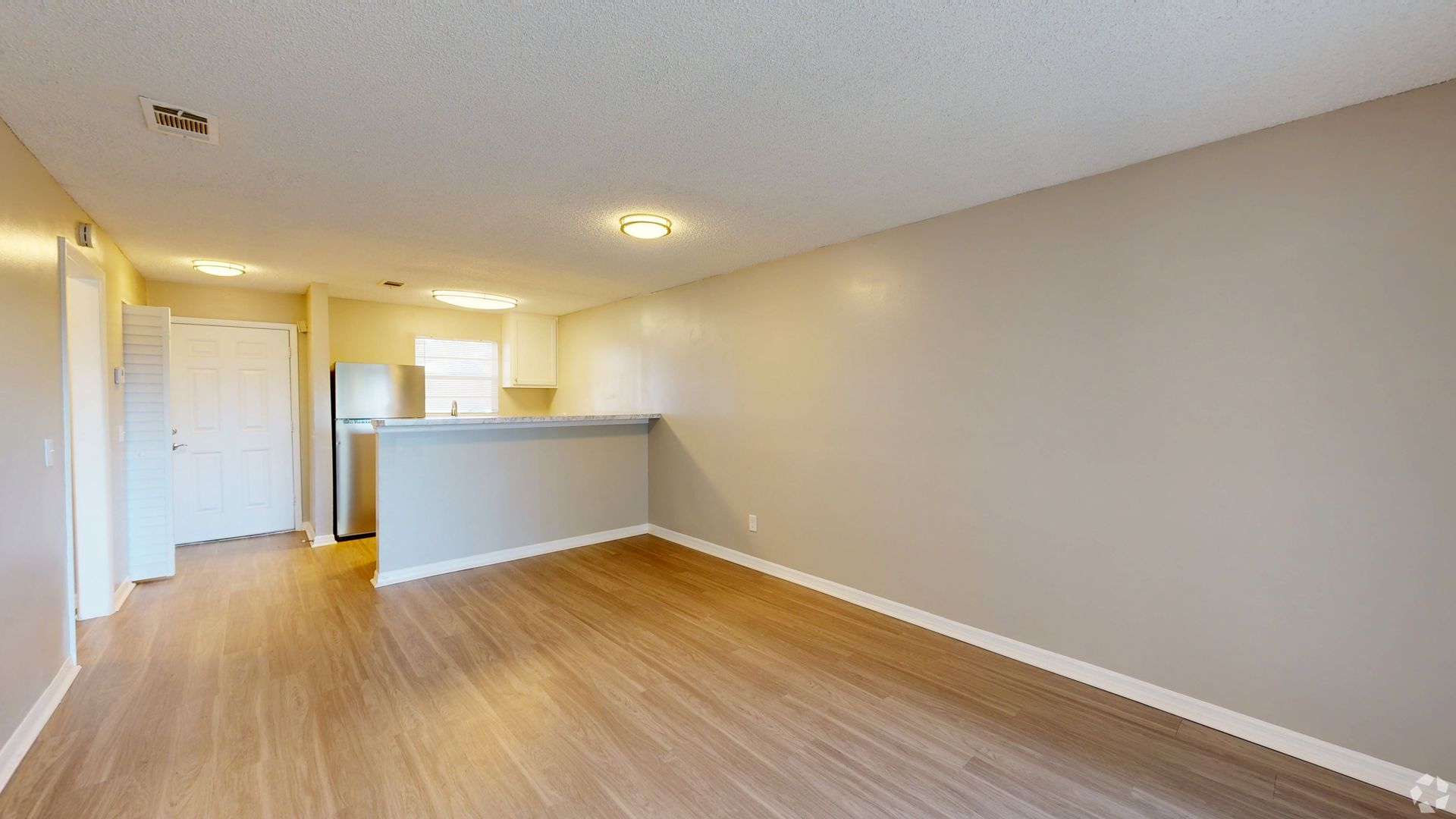 Empty, light-filled room with wood-look flooring and beige walls. Includes a kitchen area with a half-wall and appliances at Caroline Village in Jacksonville, FL.