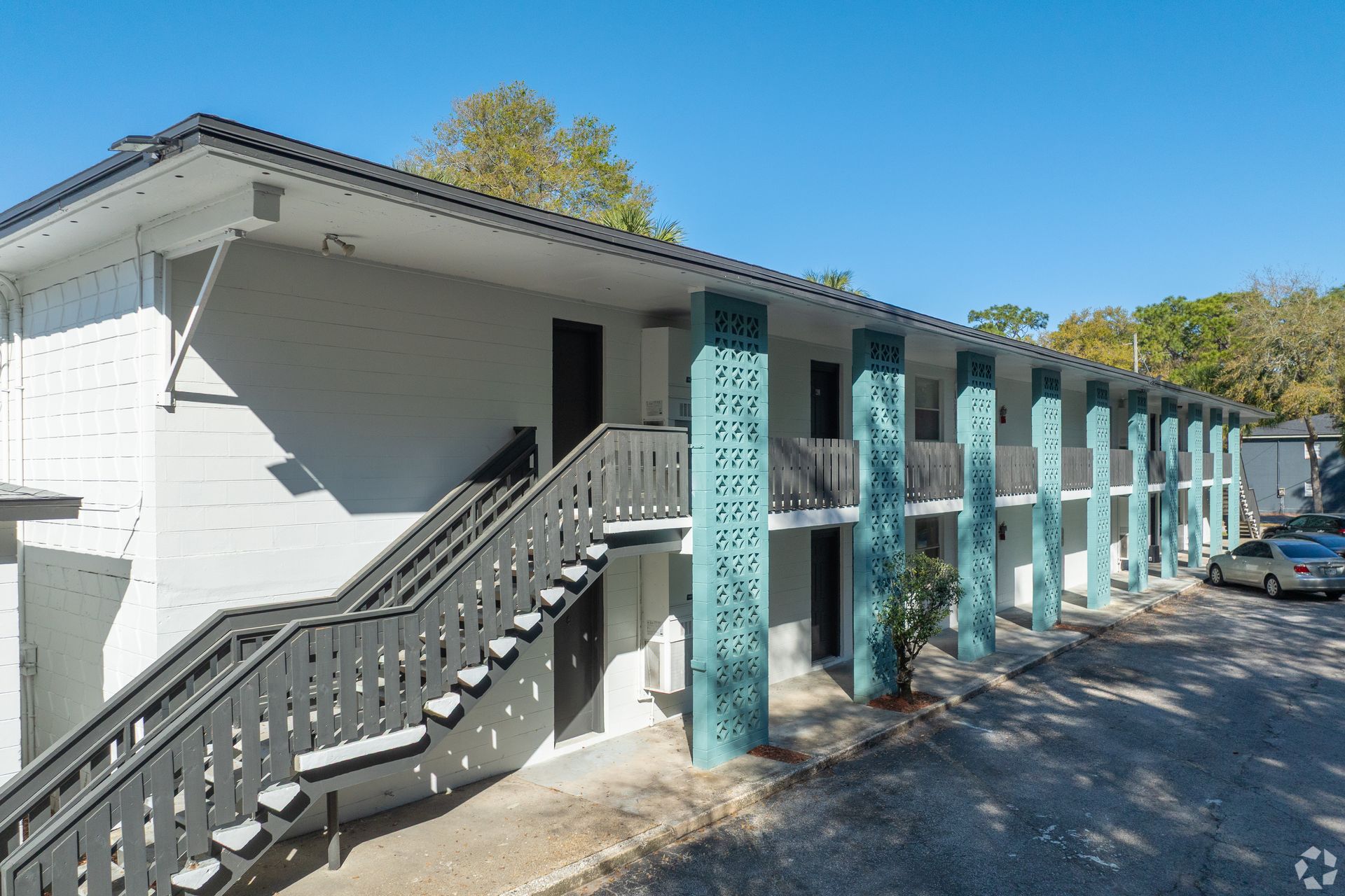Two-story apartment building with aqua columns, gray stairs, and black doors. Cars parked nearby, sunny day at Caroline Village in Jacksonville, FL.