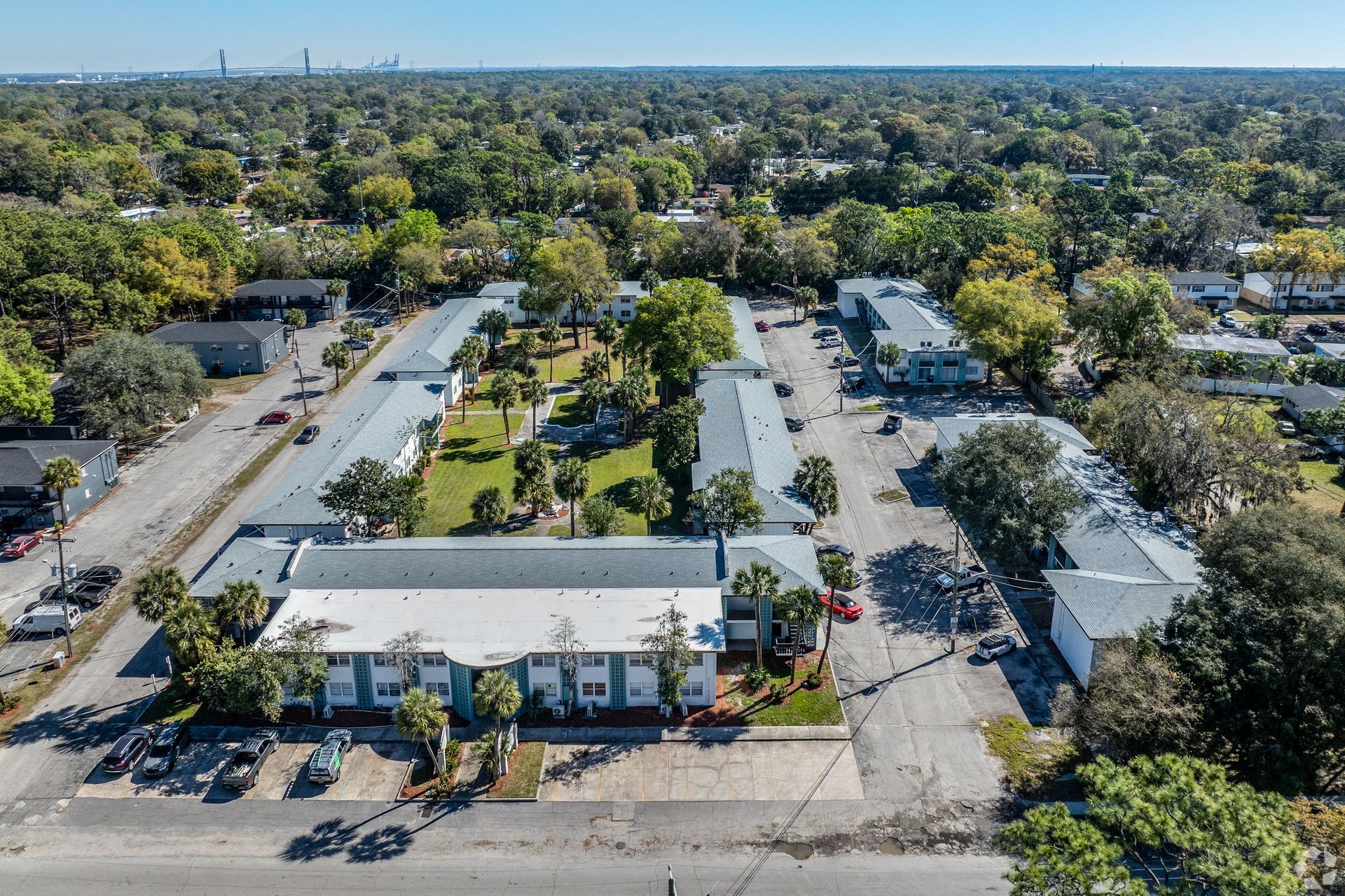 Aerial view of a long, white apartment complex with a central green space, trees, and cars in a suburban area at Caroline Village in Jacksonville, FL.