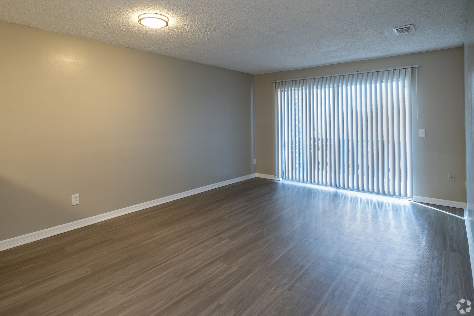 Empty room with wood-look floor, sliding glass door with blinds, beige walls, and a ceiling light at Caroline Village in Jacksonville, FL.