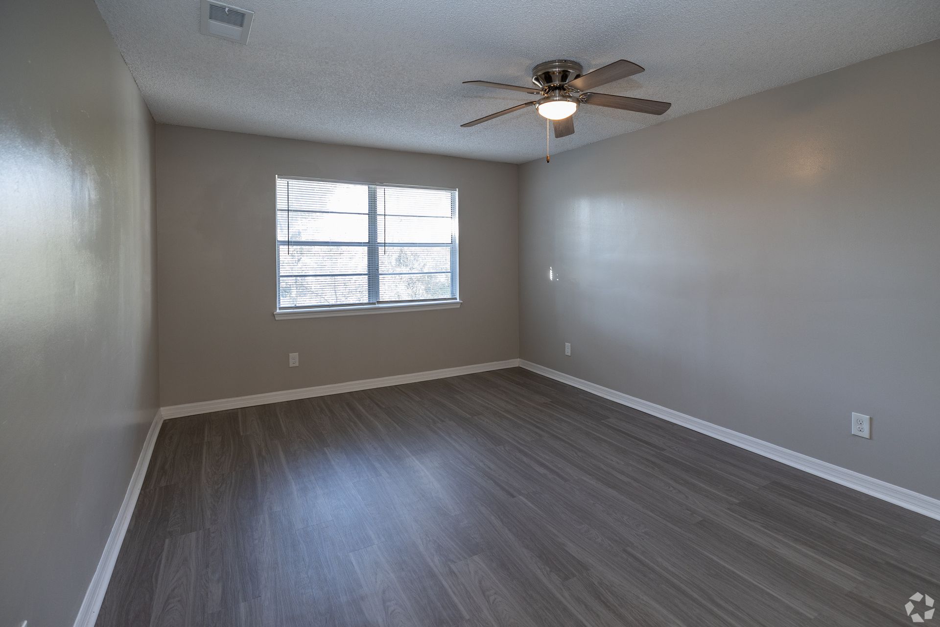 Empty room with wood-look flooring, gray walls, ceiling fan, and window with blinds at Caroline Village in Jacksonville, FL.