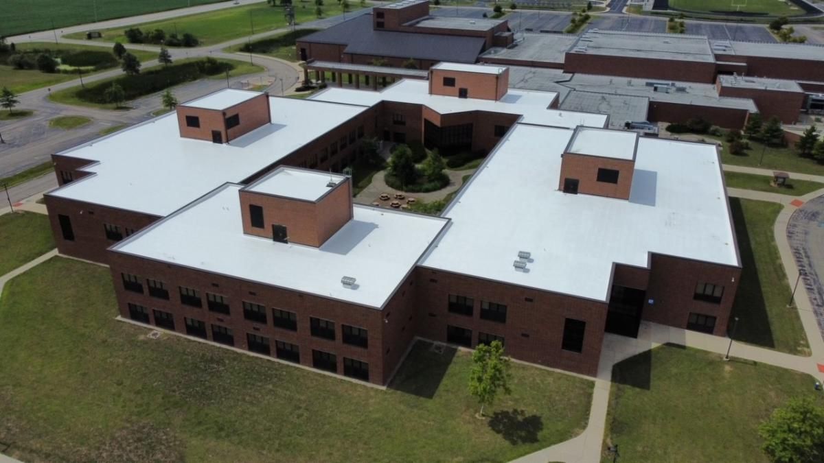 Aerial view of a large brick building with white roofs and rooftop structures, surrounded by grass and walkways