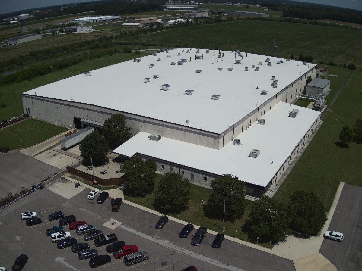Aerial view of a large white industrial building with parking lot and surrounding green fields.